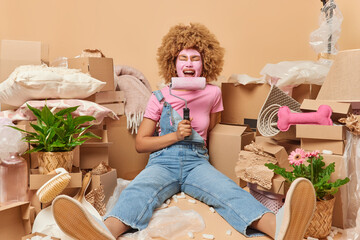 Emotional woman dressed in t shirt denim overalls and shoes holds paint roller going to repair her new house and refurbish walls in room poses on floor against cardboard boxes. Moving and improvement
