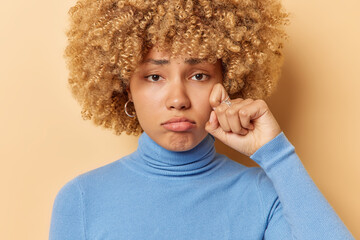 Photo of dejected frustrated woman wipes tears feels very upset after hearing bad news watches melodrama recalls unhappy memory wears blue poloneck poses against beige background. Negative emotions