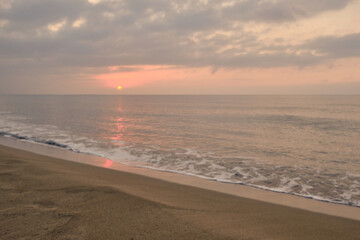 Rising sun and its reflection in sea in early morning. Cloudy sunrise at deserted sandy beach. Slight morning surf waves