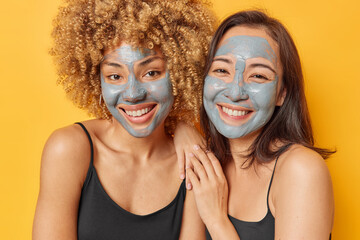 Positive mixed race women smile toothily apply nourishing clay masks for reducing fine lines and blackheads stand next to each other isolated over yellow background. Beauty and skin care concept