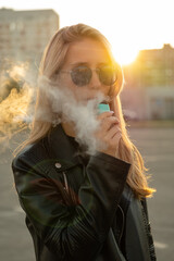 Young woman with sunglasses smoke vape pod system on a city street against the background of buildings. Female inhales and exhales vapor of electronic cigarette.