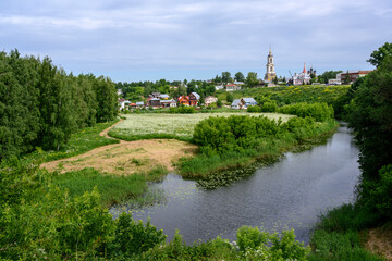Fototapeta na wymiar View of the pond, blooming meadow, forest, bell tower, churches and cottages in summer in Suzdal, Russia