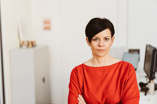 Businesswoman stands with folded arms in the office and looks thoughtfully to the camera
