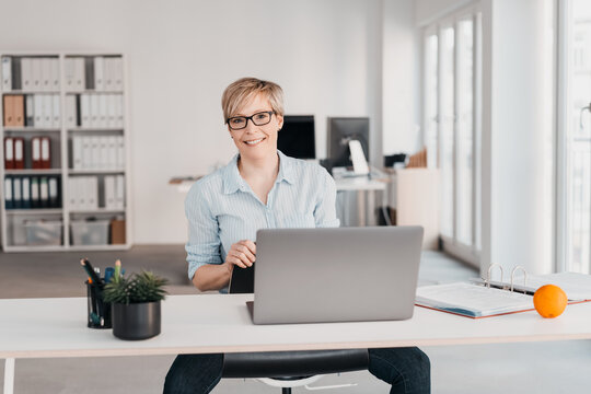 Businesswoman sitting at desk behind laptop and looking into camera with smile