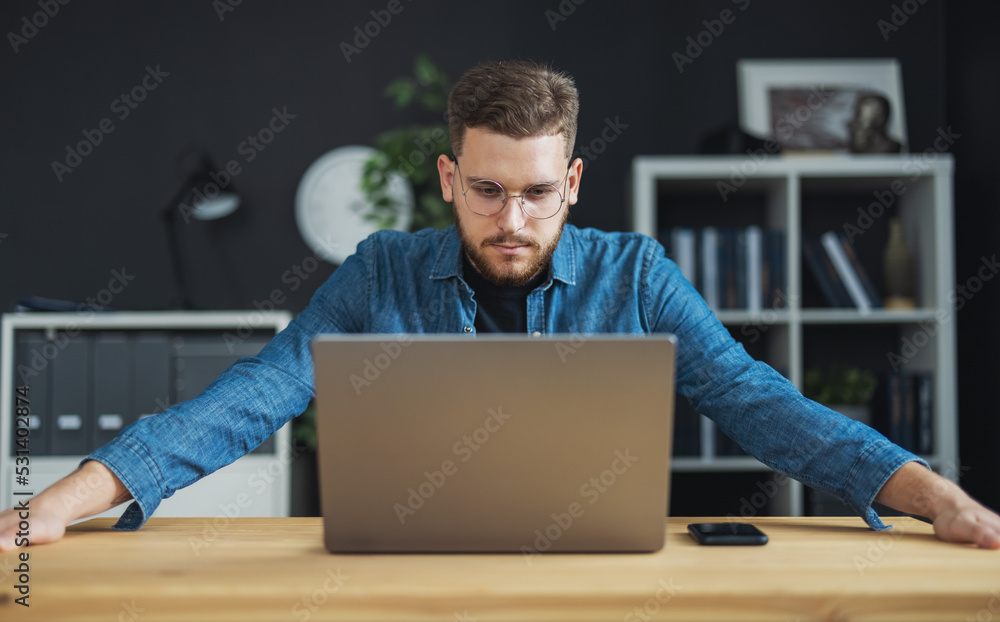 Poster confident young man in eyewear focused on laptop screen, bearded coder thoughts