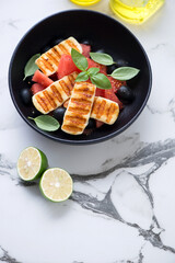 Black bowl with grilled halloumi, watermelon and olives salad, elevated view on a white marble background, vertical shot with space