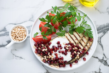 Grilled eggplant with pomegranate seeds, yogurt, tomatoes and fresh parsley, elevated view on a white marble background, horizontal shot
