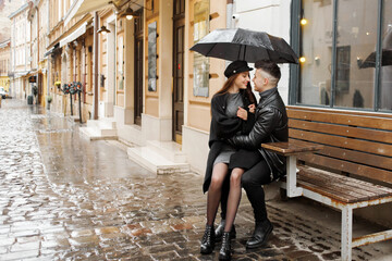 Young couple sitting near a cafe on a bench under an umbrella in rainy weather. Concept of love,...