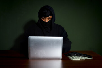 Criminal man in black hood with his gun and money looking at laptop computer at desk, mask thief robber committing committed cyber crimes online.