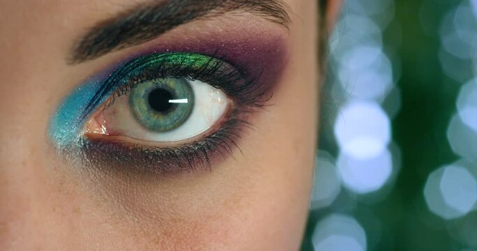 Woman open eye, with colorful makeup on eyelid and sculpted eyebrow in macro view. Model blue pupil dilate, in close up of bright color eyeshadow and long lashes in portrait against bokeh backdrop.