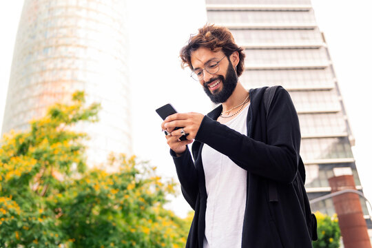 stylish young man with beard smiling happy using a mobile phone in the city, concept of urban lifestyle and technology of communication, copy space for text