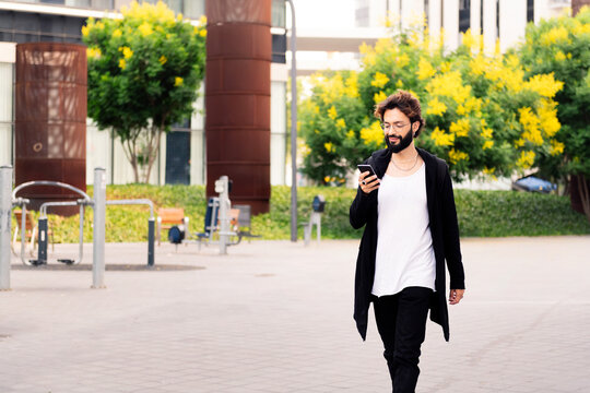 stylish young man with beard walking happy using a mobile phone in the city, concept of urban lifestyle and technology of communication, copy space for text