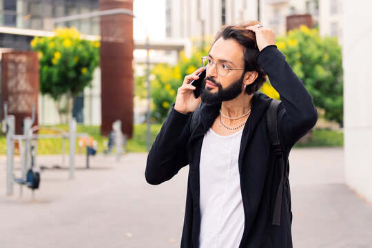 stylish young man with beard talking worried on mobile phone walking outdoors, concept of urban lifestyle and technology of communication, copy space for text