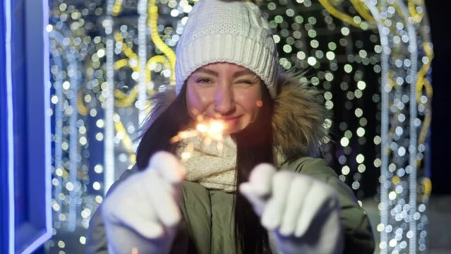 Happy woman with sparkling lights. Young female waving sparklers. Christmas lights at background. Holiday concept. Sparkling lights bengal fires. 4K, UHD