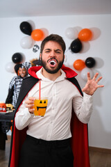 Young man dressed as vampire with a cocktail at costume Halloween party in a house.