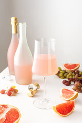 A glass of rose sparkling wine and bottles of alcoholic grapefruit wine on a white table surrounded by slices of grapefruit and grapes