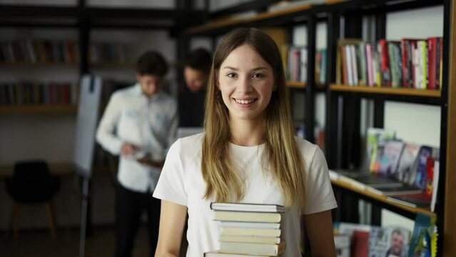 Student girl walks through library with stack of books in hands, shelves with book, front view. Young woman hold books at hands walks through library