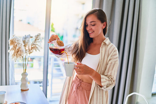 Happy young cute joyful smiling satisfied woman pouring hot tea into cup from glass teapot at home - Powered by Adobe