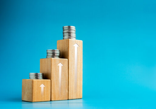 Shining rise up arrow on wooden cube blocks top with coins stacked, bar graph chart steps on blue background with space, investment, income, inflation, business growth, economic improvement concepts.