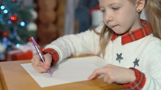 Cute little girl draws a kitten with a pencil on paper at home. Portrait of a child in a room on the background of a Christmas tree.