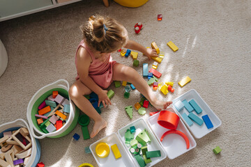 A little girl playing with colorful wooden blocks on floor in nursery. Educational games for Colors sorting. Boxes with red, yellow, green and blue toys. Developing Montessori toddlers activities.