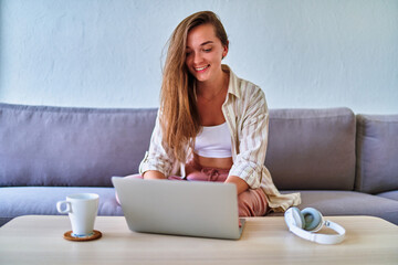 Young happy smiling casual smart modern cute satisfied girl sitting on the sofa and working online at laptop at home