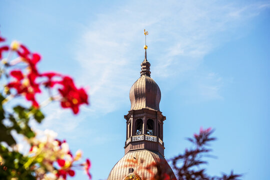 A church with a brown wall and a pointed metal tip in the middle of the city center with pink flowers - Powered by Adobe