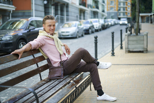 Handsome man sits on the bench with legs crossed spend time in urban city near his office or home, wearing pink shirt and shoulder bag. Freelancer man off work