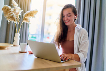 Young happy joyful smart modern smiling cute satisfied girl using computer at home interior