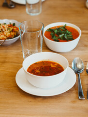 Table set for dinner. On wooden table is a bowl with tomato soup, salad bowl and basil sauce