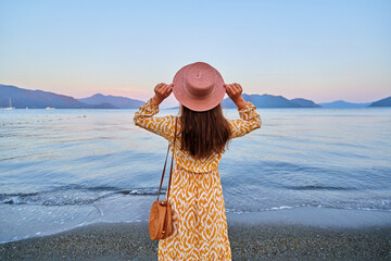 Back of serene calm pensive girl traveler wearing hat and dress stands alone against the backdrop of the sea and mountains