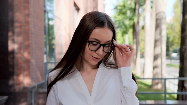 Close up portrait attractive serious confident business woman, employee looking at camera. Pretty female banker lawyer worker stand near office place building. Confident lady touches glasses with hand