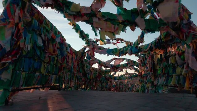 smooth flight of camera through the arched tunnel of colorful silk flags bringing good luck and the sun peeping through them in a buddhist temple against the blue sky during the dusk or dawn