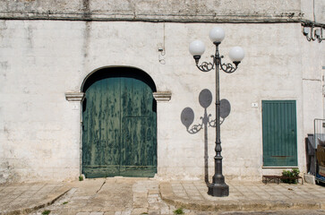 Un lampione getta la sua ombra vicino a un portone verde sul muro bianco di un paese in Salento, in Puglia