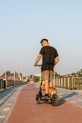 European man is riding on electric scooter with his small dog. Walking with dog on empty street of city at sunny day.