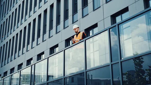 Confident architect engineer walking on the terrace of a modern building using mobile phone, reading message answering outdoors wearing in a safety helmet at a building construction site. 