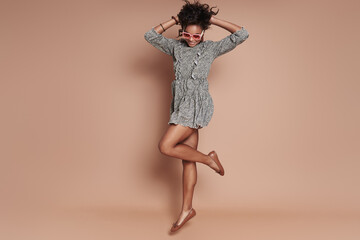 Full length of beautiful African woman in funky eyeglasses jumping against brown background