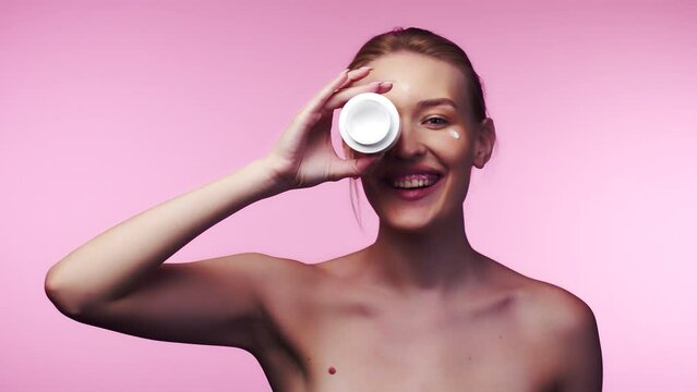 Happy beautiful young caucasian woman apply cream on face laugh looking at camera holding cream jar to her eye. Studio, advertisement, facial skin care moisture treatment hydration concept