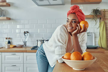Thoughtful African woman in traditional headwear holding orange while standing at the kitchen