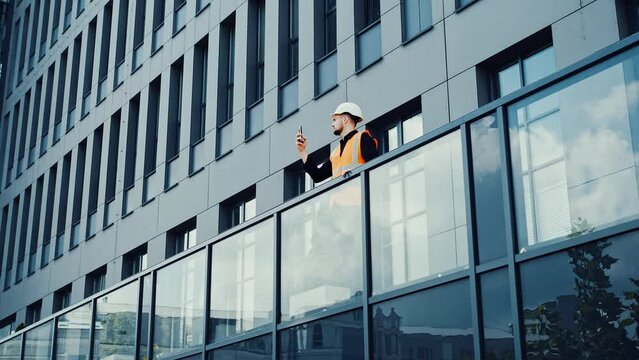 Professional architect engineer walking on the terrace of a modern building using mobile phone shooting video makes a photo outdoors wearing in a safety helmet at a building construction site.
