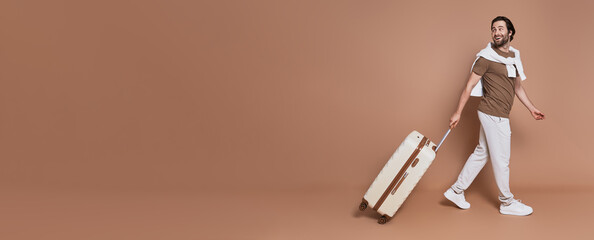 Full length of handsome young man with suitcase walking against brown background