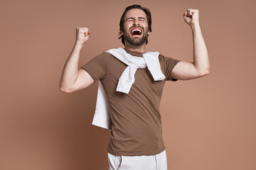 Excited young man keeping eyes closed and gesturing against brown background