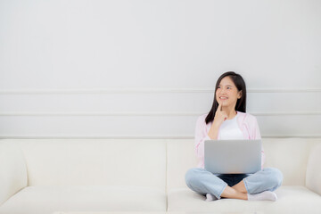 Young asian woman working on laptop computer sitting on sofa in the living room at home, female is...