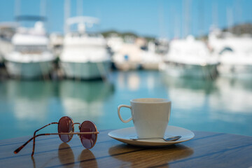 White cup with saucer, sunglasses on a table on the background of blurred boat harbor. Summer...