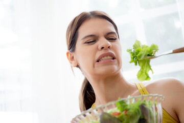 Young caucasian woman sitting on sofa eating vegetable salad while feeling dislike and expression...