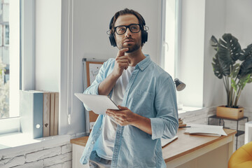 Fototapeta premium Thoughtful young man in headphones holding digital tablet while standing in the office