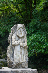 Fototapeta na wymiar Stone statue standing among green trees in park symbolises old forest dwellers. Vintage sculpture attracts tourists attention among plants