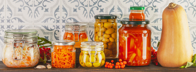 Fototapeta na wymiar Food banner with autumn seasonal pickled or fermented vegetables and mushrooms in jars placed in row on wooden bord on tile wall background. Home food preserving or canning. Vegetarian and vegan foods