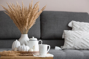 Still-life. Dried pampas grass in a vase, white ceramic pumpkins, a teapot, a cup and pumpkin-shaped candles on a coffee table in the home interior of the living room. Cozy autumn concept.