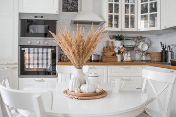 Still-life. Dried pampas grass in a vase, white ceramic pumpkins, a teapot and pumpkin-shaped candles on a white table in the interior of a Scandinavian-style home kitchen. Cozy autumn concept.